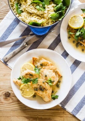 Chicken piccata with white wine and capers with lemon on a white plate, with a pot of cracked wheat and spinach in the background.