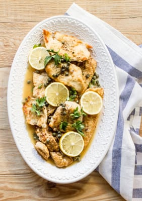 Chicken piccata with white wine and capers, sliced lemons, on an oval-shaped white serving platter on a wooden surface.