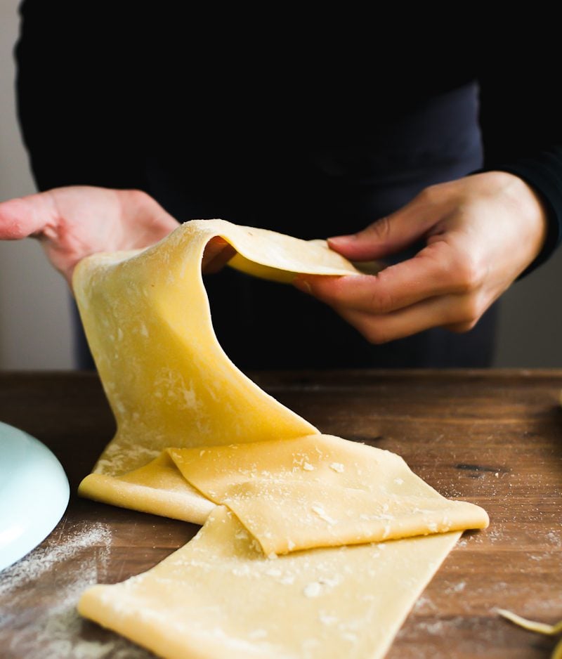 Two hands holding up a long, silky rolled sheet of fresh pasta dough.