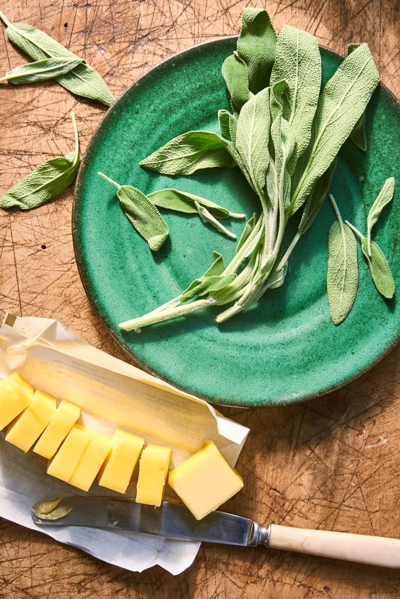 Green plate with sage leaves on top, and a stick of butter cut into pats on a cutting board with a knife.