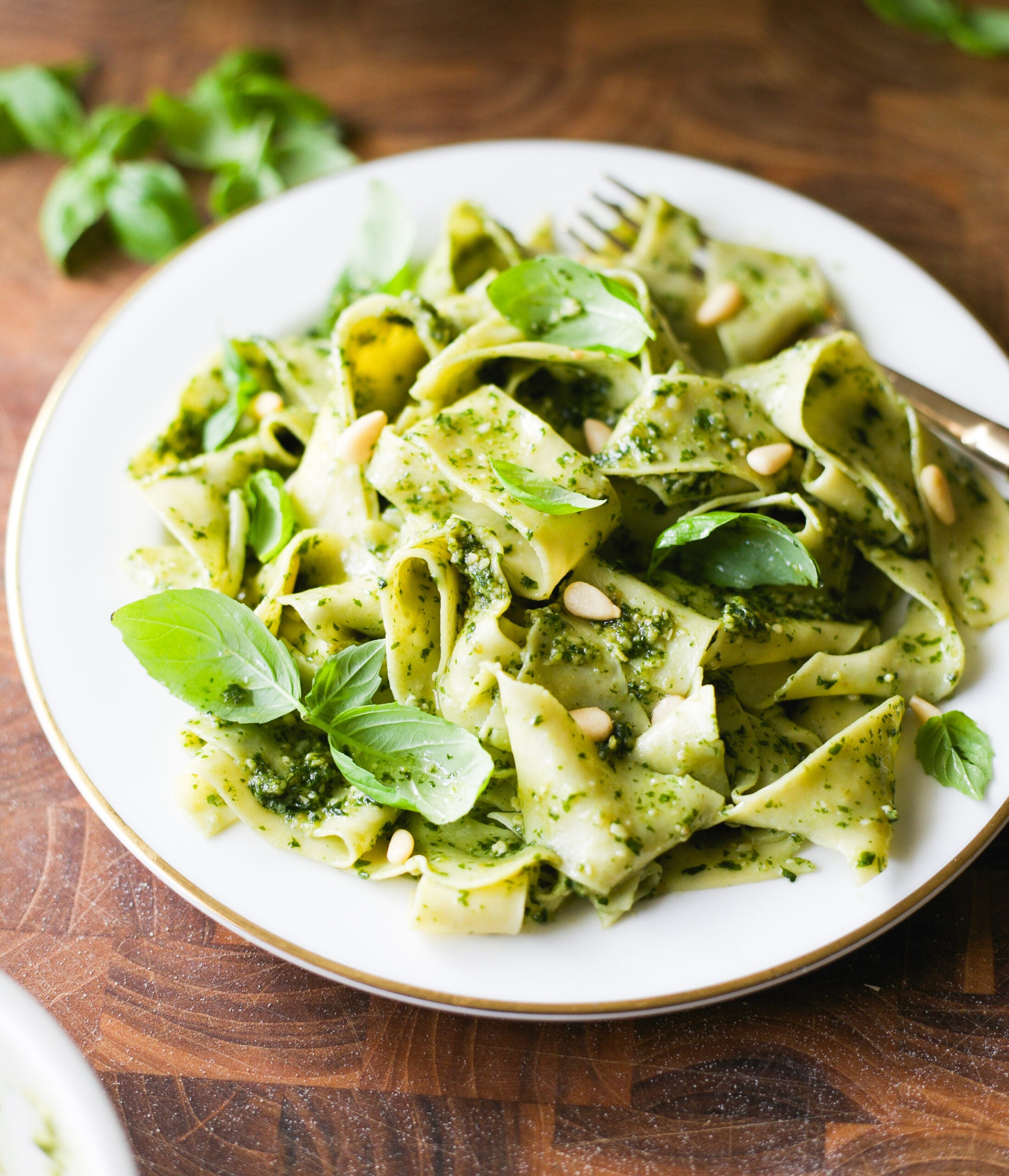 Pappardelle pesto on a white plate, topped with fresh basil leaves and pine nuts.