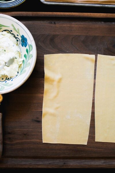 Fresh homemade pasta sheet on a dark wooden surface with ricotta cheese filling in a bowl.