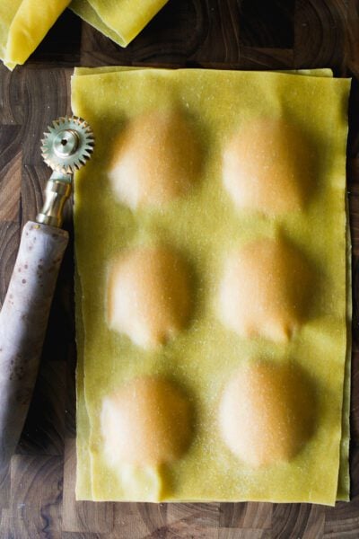 Butternut squash filling for ravioli tucked inside a fresh pasta sheet, set on a wooden surface with a fluted pastry wheel.