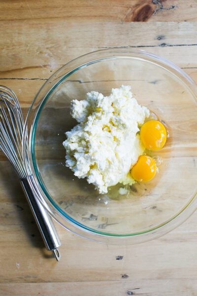 Ricotta and eggs in a glass mixing bowl with a whisk on the side