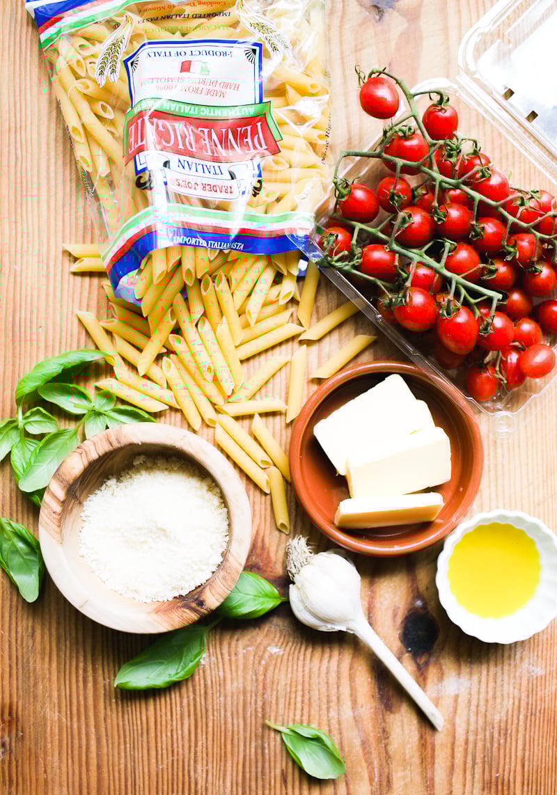 Penne pasta recipe ingredients: penne, cherry tomatoes, fresh basil, butter, oil, garlic and Parmesan cheese on a wooden surface.