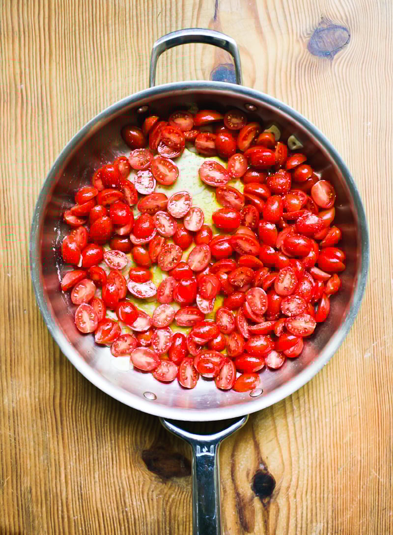 Melted oil, sliced garlic and halved cherry tomatoes in a stainless steel pan on a wooden surface.