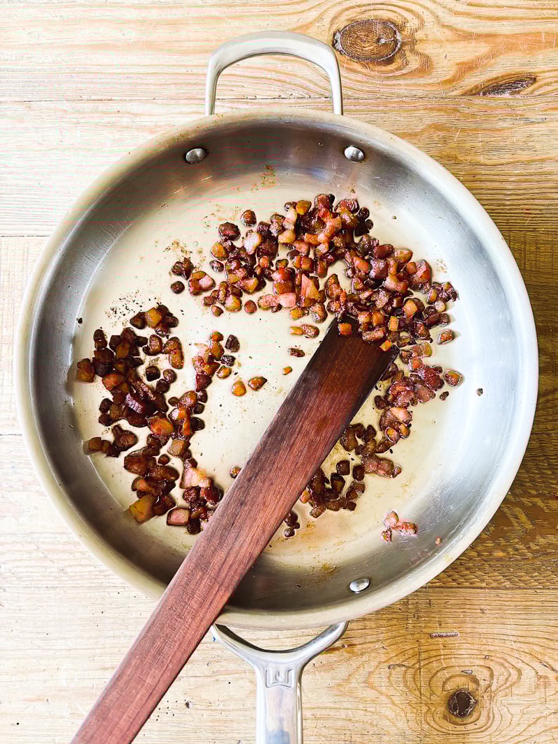 Sauteed, cubed guanciale, golden and crisp, in a large stainless steel pan with a wooden spoon.