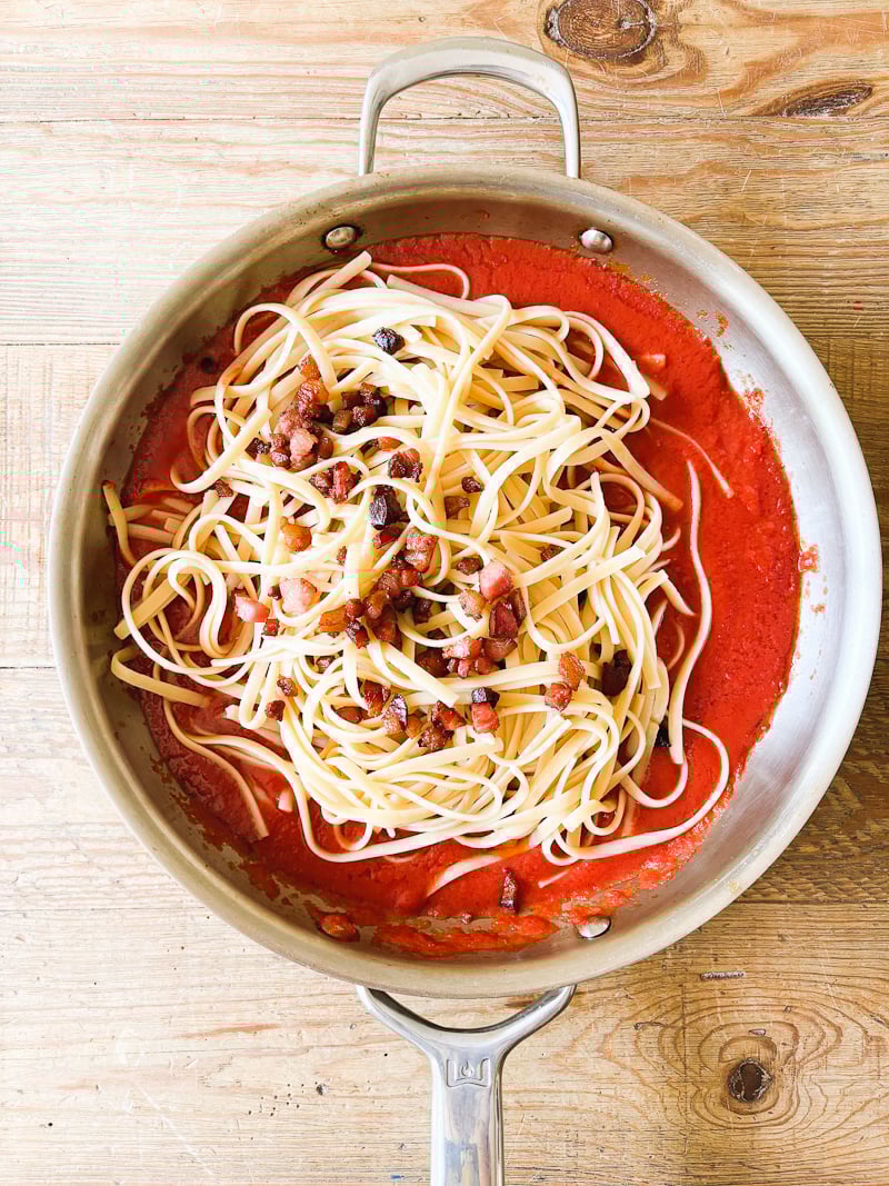 Cooked pasta, guanciale, and tomato passata in a large stainless steel pan.