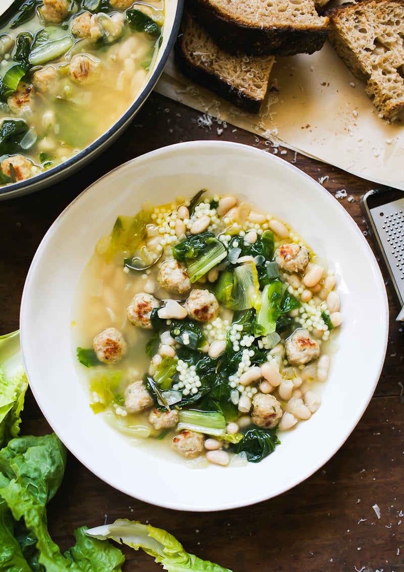 Italian wedding soup with escarole, tiny chicken meatballs, and white beans in a white bowl with crusty sourdough bread