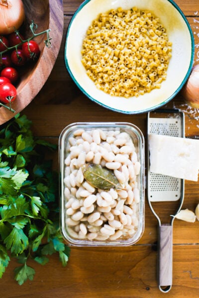Home-cooked white beans in a glass container on a rustic wooden surface with pasta e fagioli ingredients