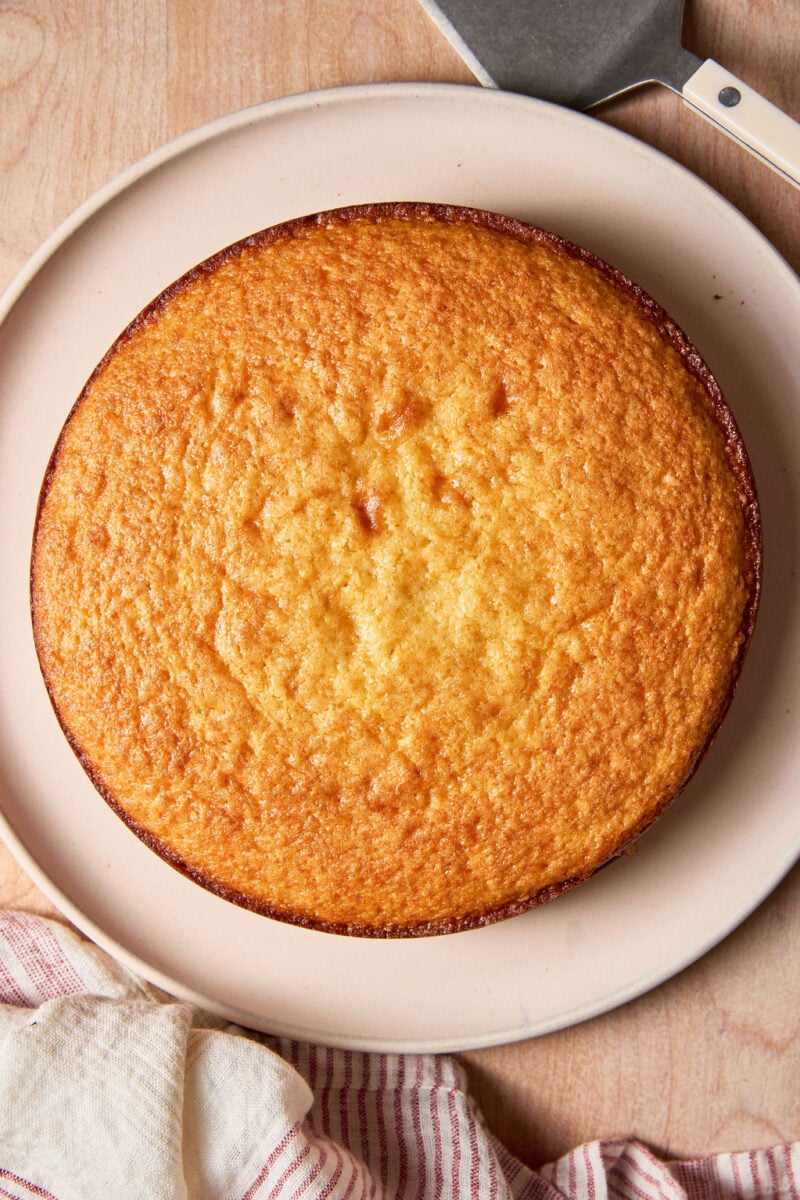 Baked sourdough French yogurt cake on a pale pink plate, set on a wooden surface with a cream and white striped linen towel.