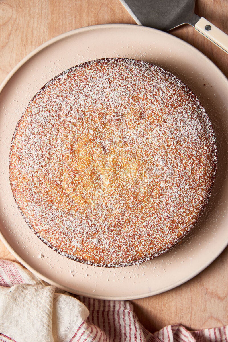 Sourdough French yogurt cake with powdered sugar on a pale pink plate, set on a wooden surface with a cream and white striped linen towel.