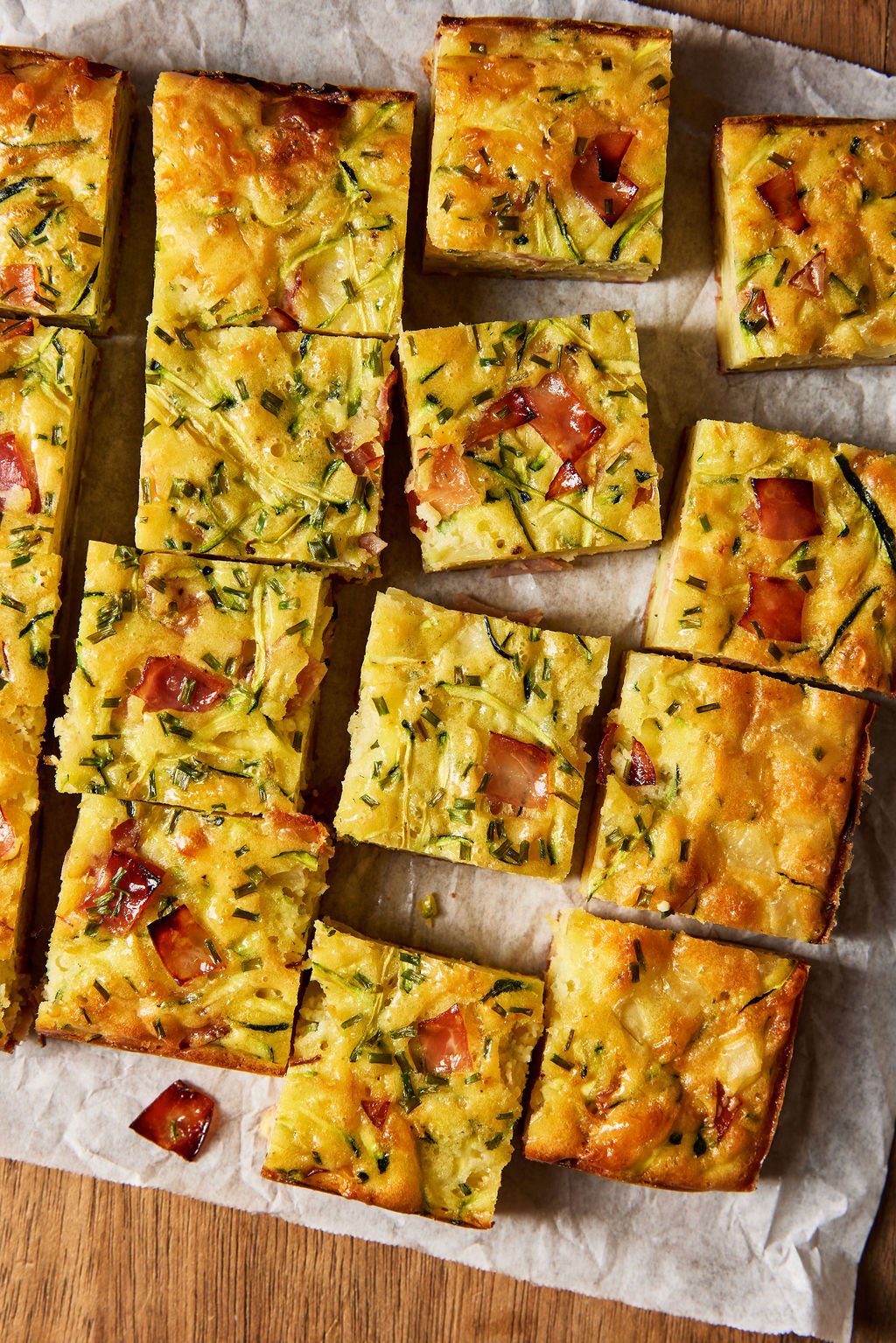 Sourdough zucchini slice made with sourdough discard, grated zucchini, cheddar cheese, ham, chives and eggs, baked and cut into squares on a parchment-lined, wooden cutting board.