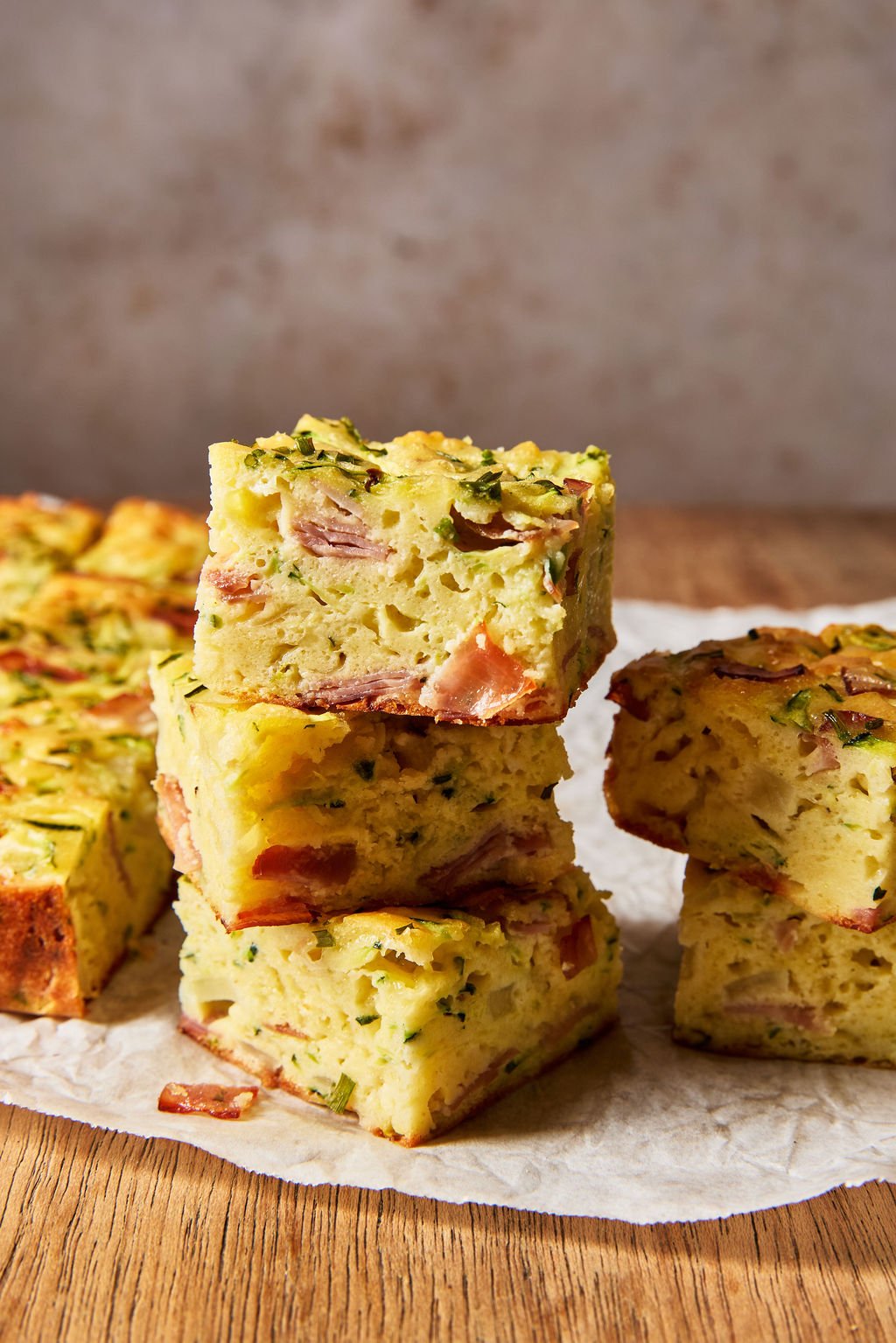 Sourdough zucchini slice made with sourdough discard, grated zucchini, cheddar cheese, ham, chives and eggs, cut into squares and stacked on a parchment-lined, wooden cutting board.