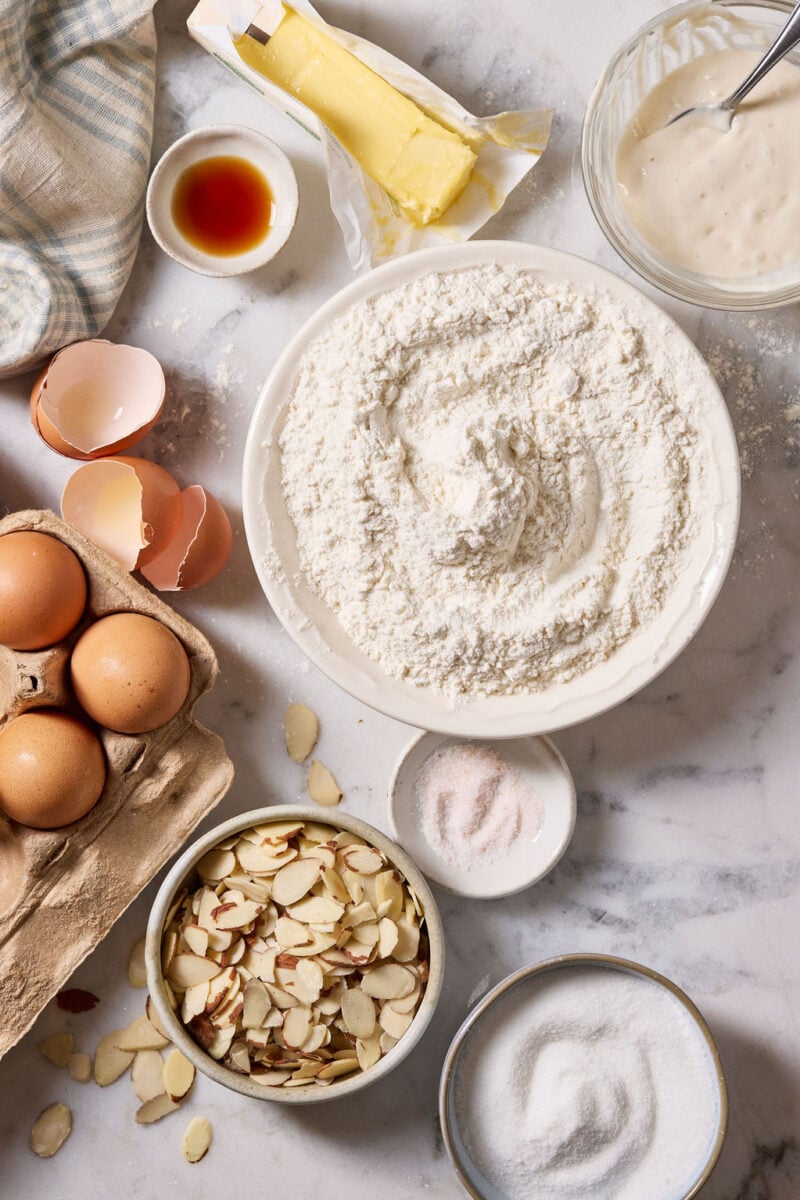Sourdough biscotti ingredients on a marble surface: bowl of all purpose flour, brown eggs, vanilla and almond extracts, softened butter, salt, sugar and sourdough starter.
