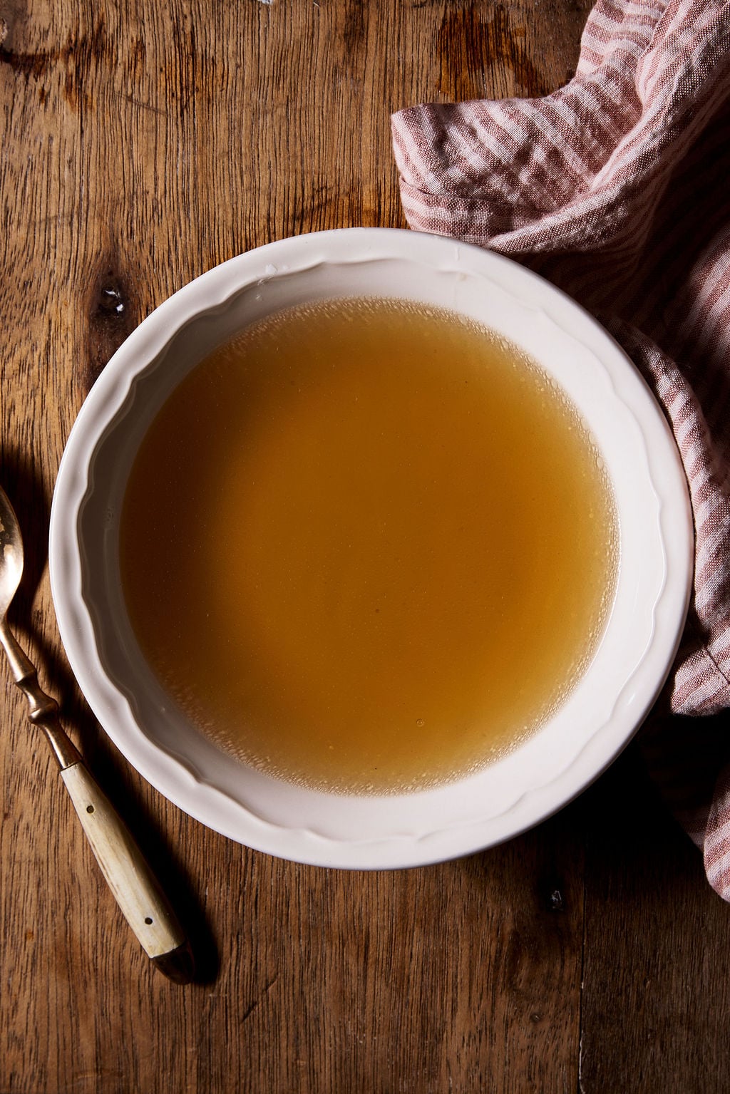Bowl of Italian chicken brodo on a wooden surface, with a spoon and red and cream striped linen tea towel off to the side.
