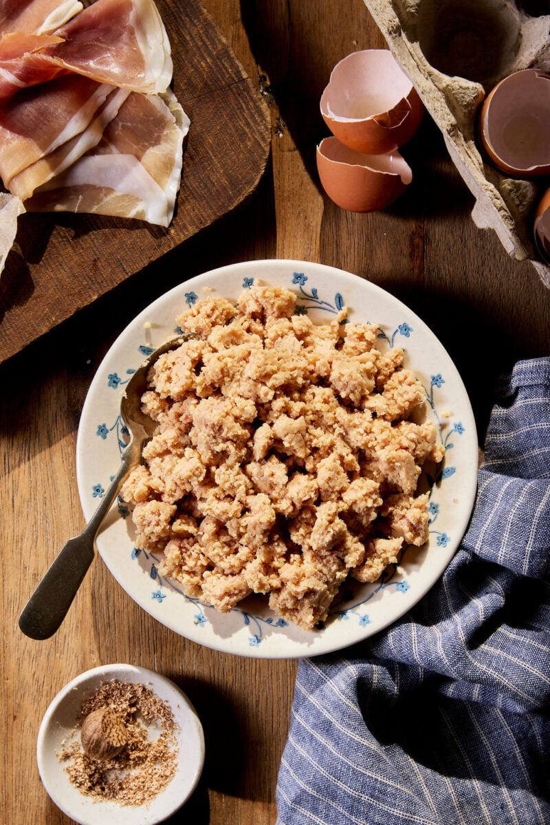 Finished, finely chopped pork filling in a white bowl on a wooden surface with egg shells and additional prepped ingredients.