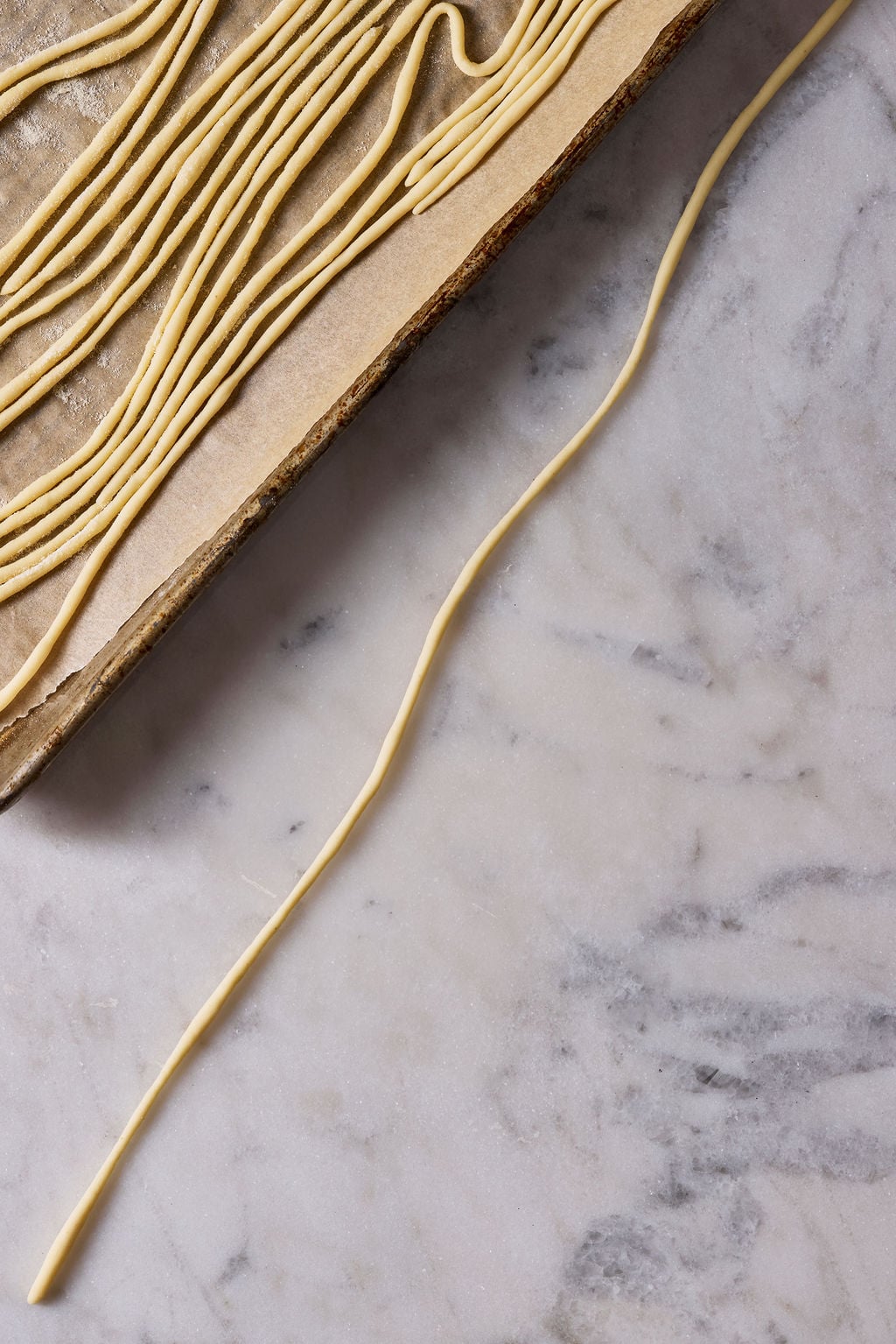 Long, sourdough pici pasta strand rolled by hand on a marble surface with a tray of finished rolled pici in the background.