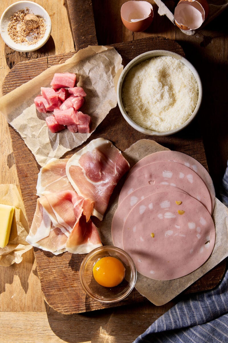 Classic pork filling for sourdough tortellini on a wooden surface: cubed, raw pork loin, prosciutto, mortadella, egg, and freshly grated nutmeg in a small white bowl.