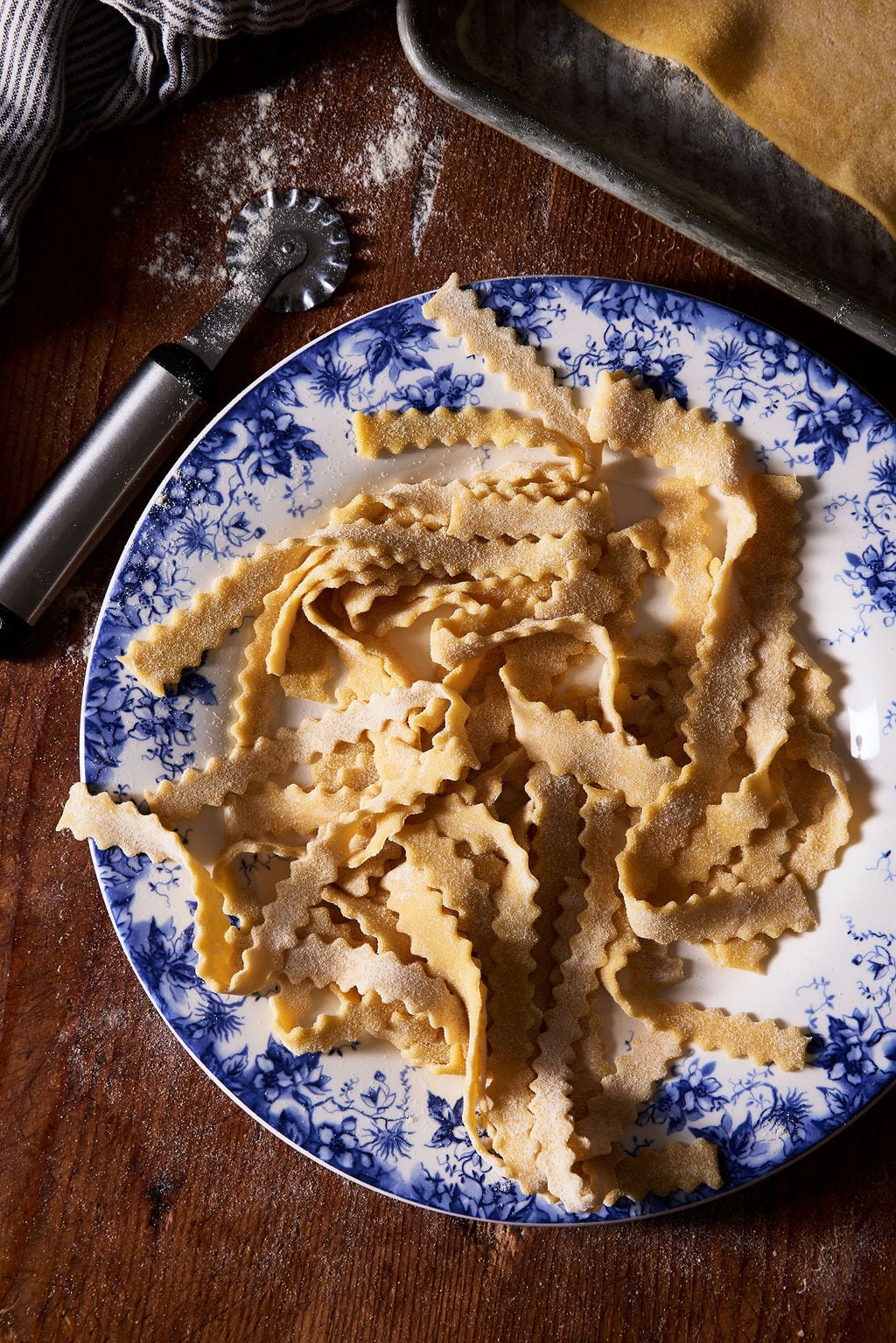 Sourdough mafaldine pasta on a blue and white plate with a fluted pastry wheel dusted with flour on a wooden surface.
