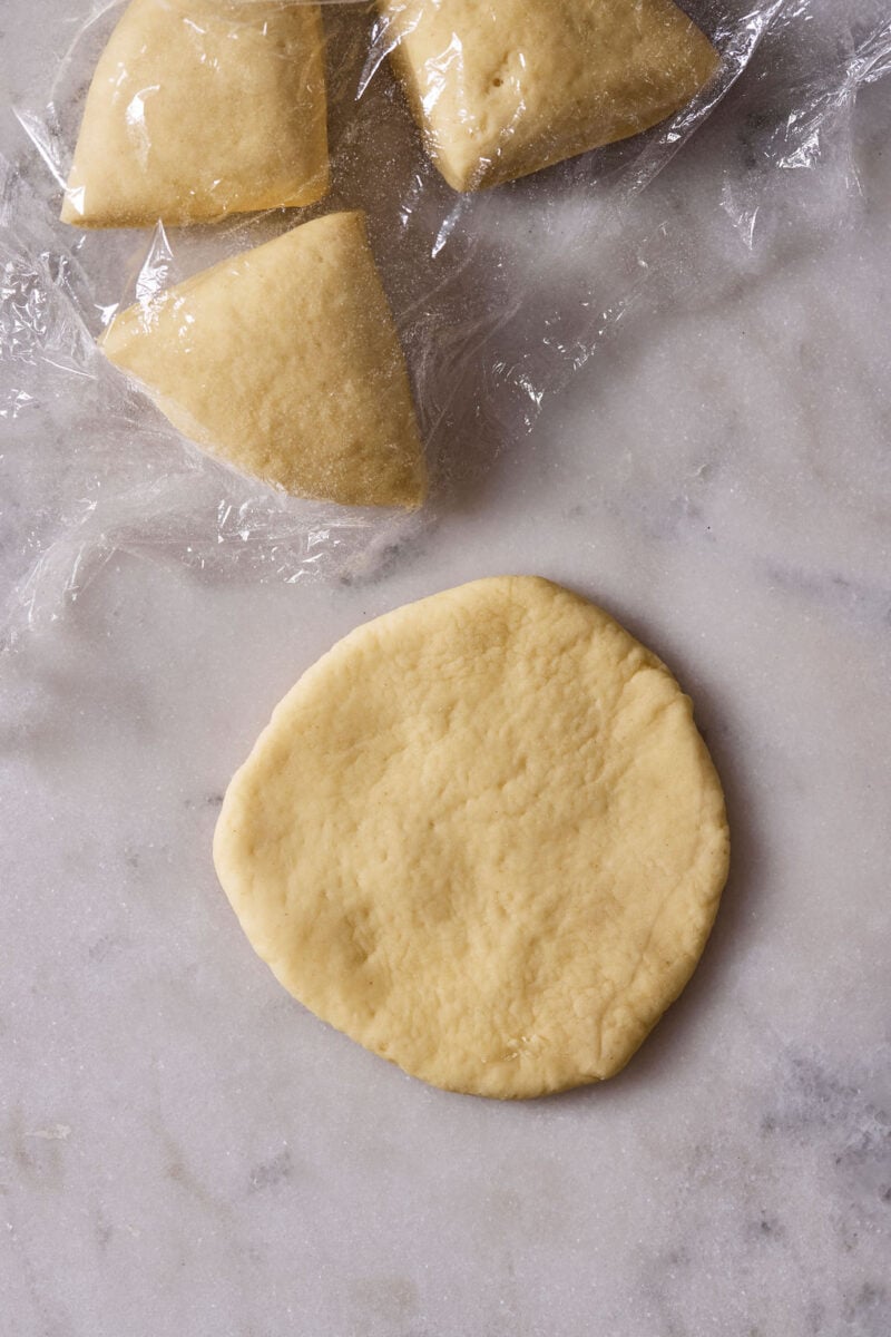 Sourdough pici pasta dough (one piece) flattened into a palm-sized disc on a marble surface, with the remaining doughs covered in plastic wrap.