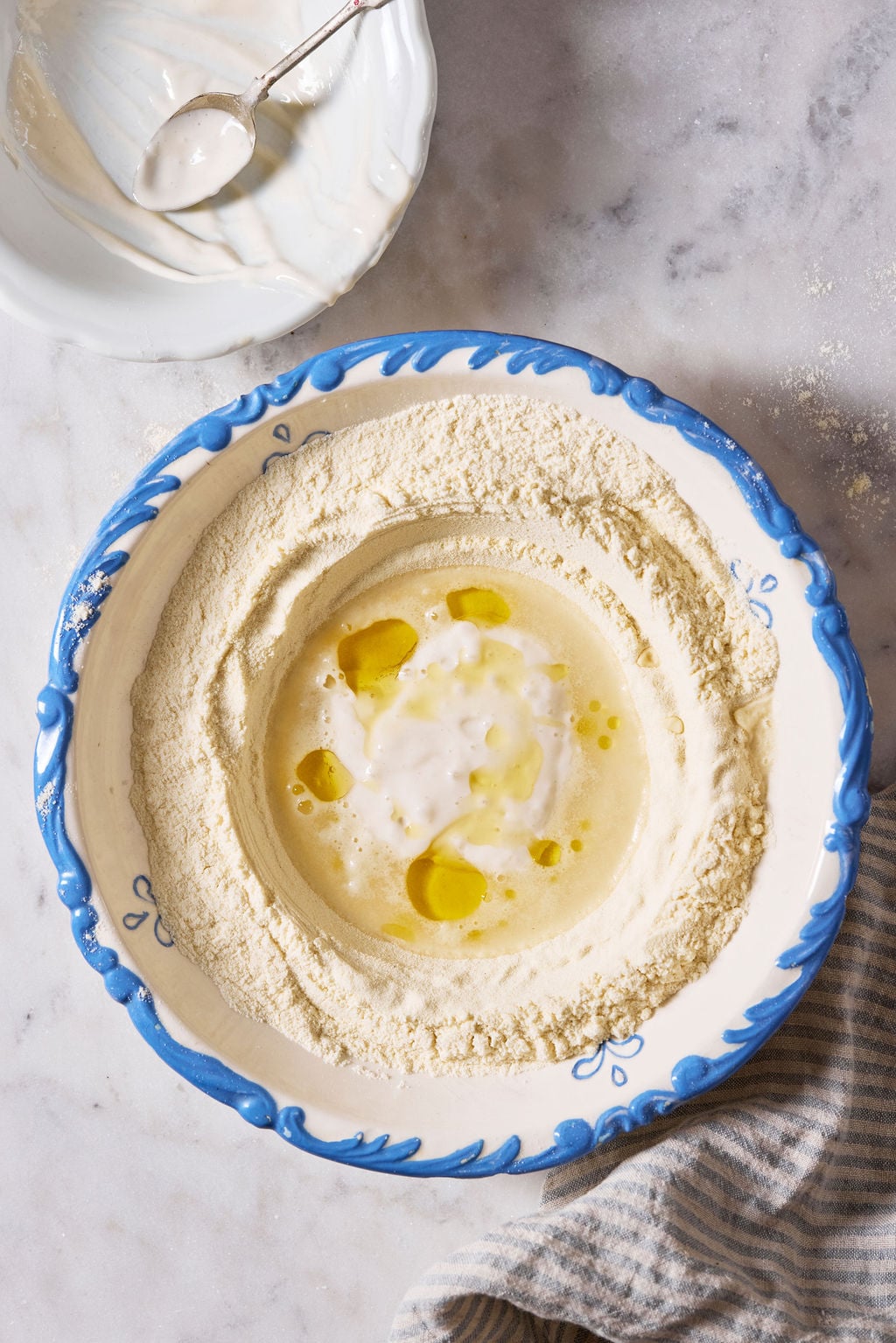 Semolina flour, warm water, sourdough starter, and olive oil in a white bowl with a blue rim.