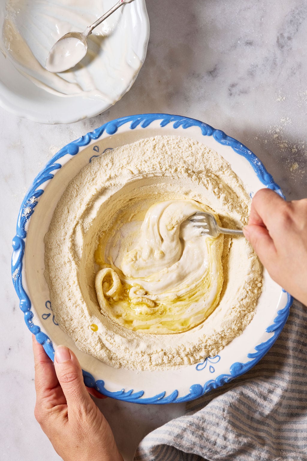 Mixing semolina flour, warm water, sourdough starter, and olive oil by hand with a fork, in a white bowl with a blue rim.