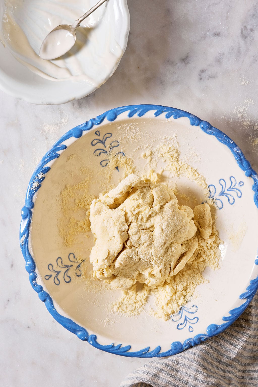 Ball of sourdough pici pasta dough in a large white bowl with a blue rim, with a small bowl of leftover sourdough starter on a marble surface.