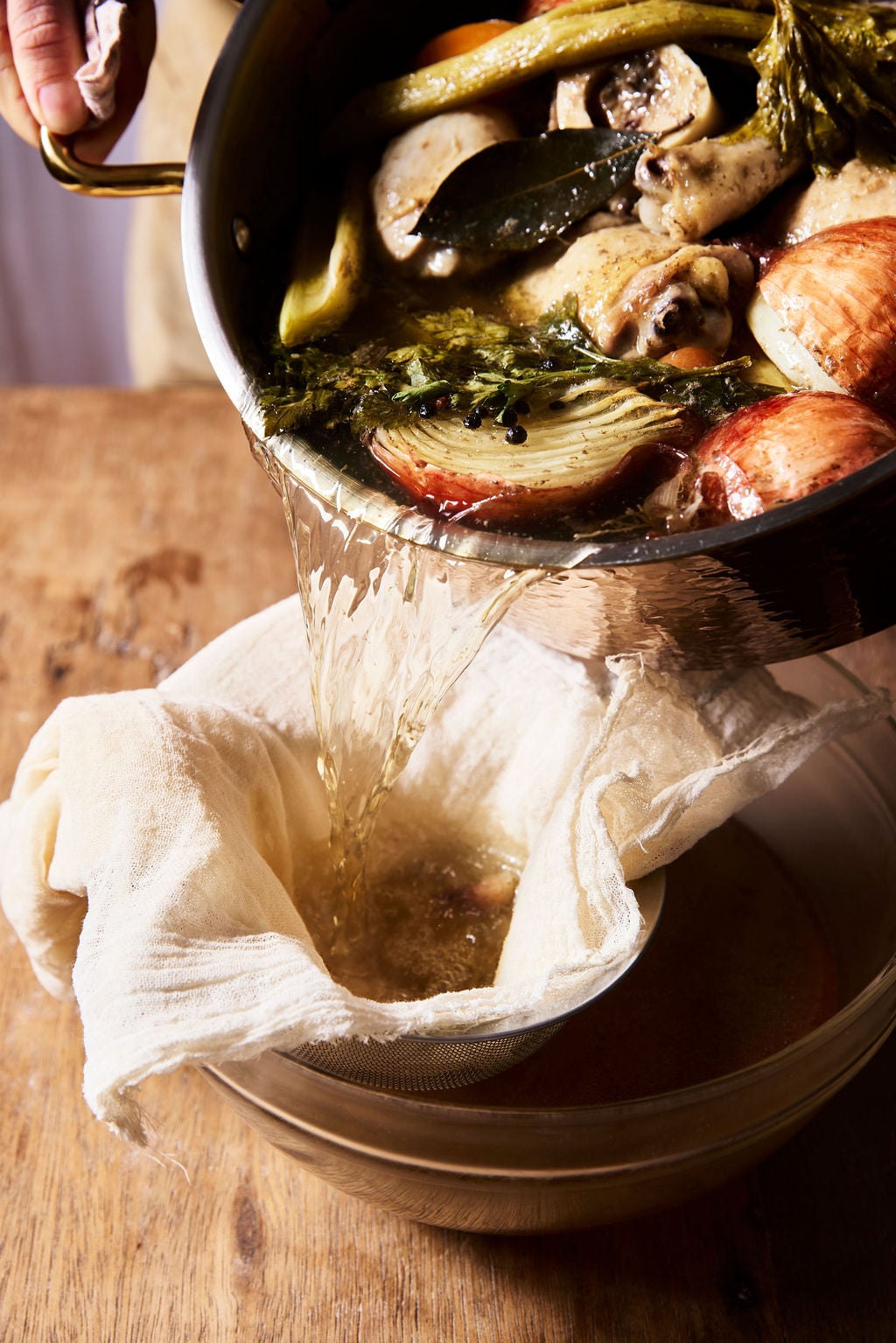 Straining Italian chicken brodo through a cheesecloth-lined strainer set over a large bowl.