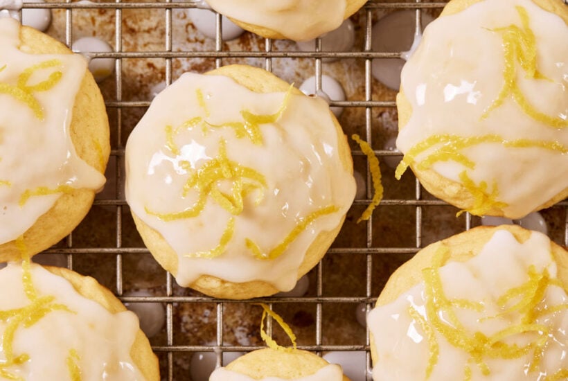 Glazed sourdough lemon drop cookies with lemon zest on a wire rack set over a sheet pan, with a metal spoon.