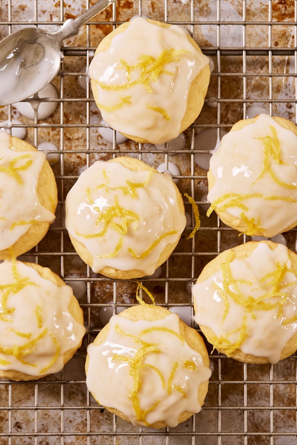 Glazed sourdough lemon drop cookies with lemon zest on a wire rack set over a sheet pan, with a metal spoon.