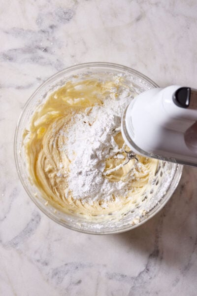Mixed cream cheese, butter and powdered sugar in a glass bowl with a hand-held electric mixer.