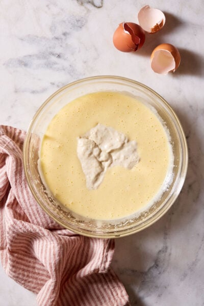 Sourdough cupcake batter with sourdough discard in a large bowl, set on a marble surface with a red-striped linen kitchen towel and egg shells.