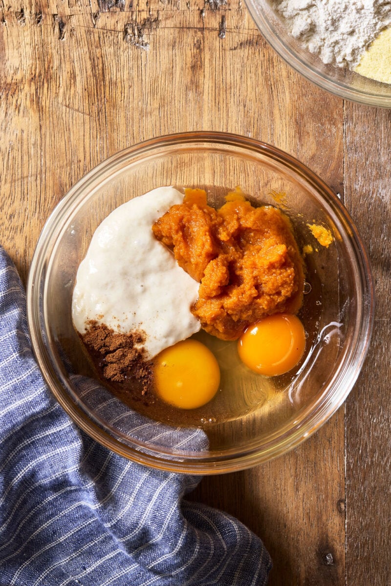 A glass bowl containing pumpkin puree, two raw eggs, sourdough starter, and a sprinkle of pumpkin spice on a wooden surface.