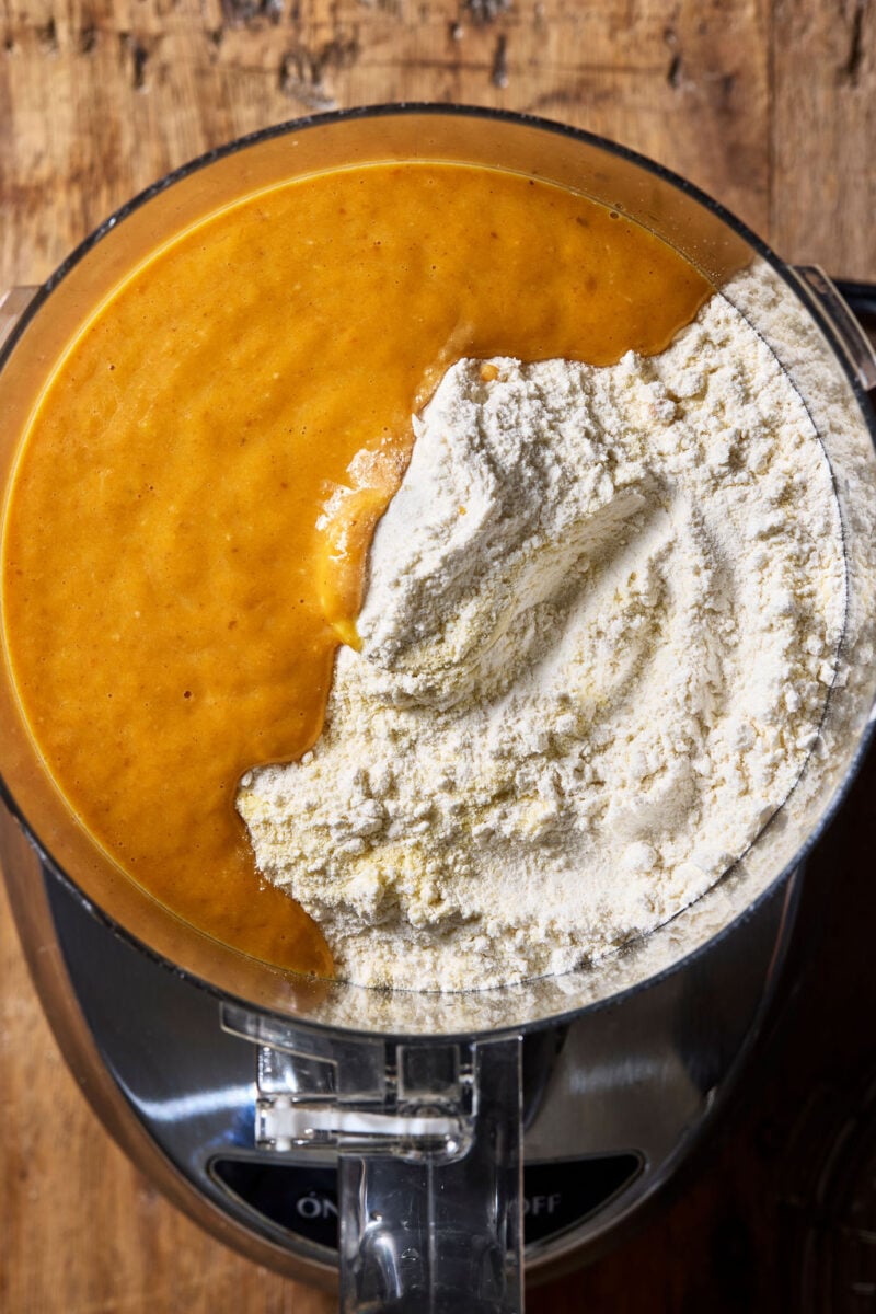 A food processor bowl containing a mixture of golden-orange pumpkin purée and a mound of white flour and semolina flour.