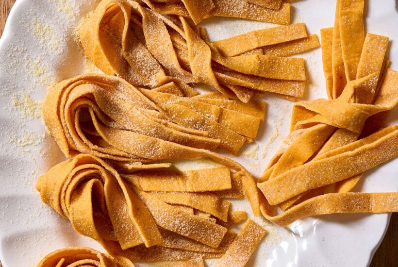 Several nests of fresh, sourdough pumpkin pasta noodles dusted with flour on a white scalloped plate.