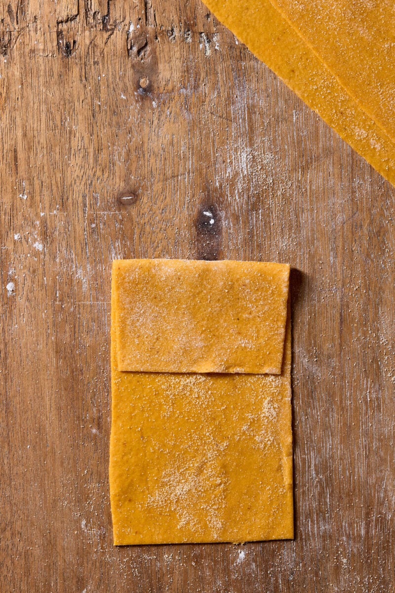 A sheet of fresh orange-hued sourdough pumpkin pasta dough folded over on a floured wooden surface.