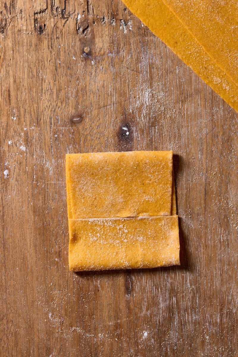 A sheet of fresh sourdough pumpkin pasta dough folded into a rectangle on a floured wooden surface.