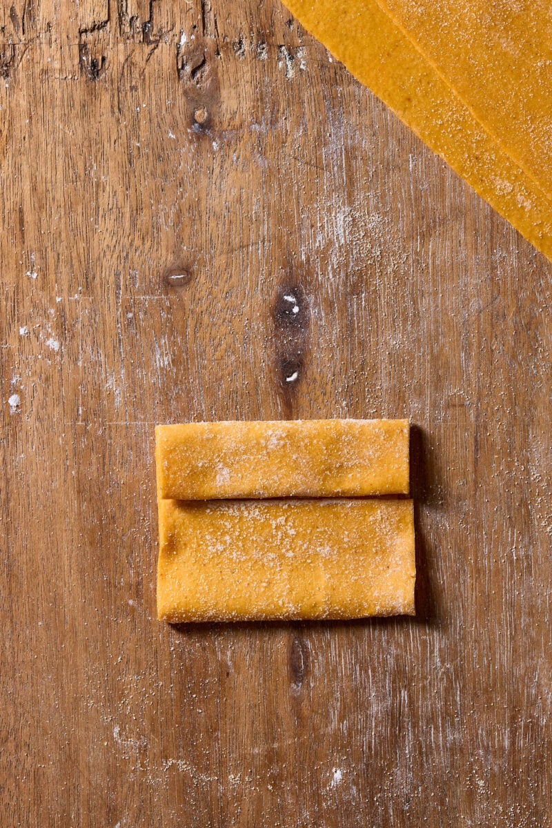 A sheet of fresh orange-hued sourdough pumpkin pasta dough folded into a rectangle on a flour-dusted wooden surface.