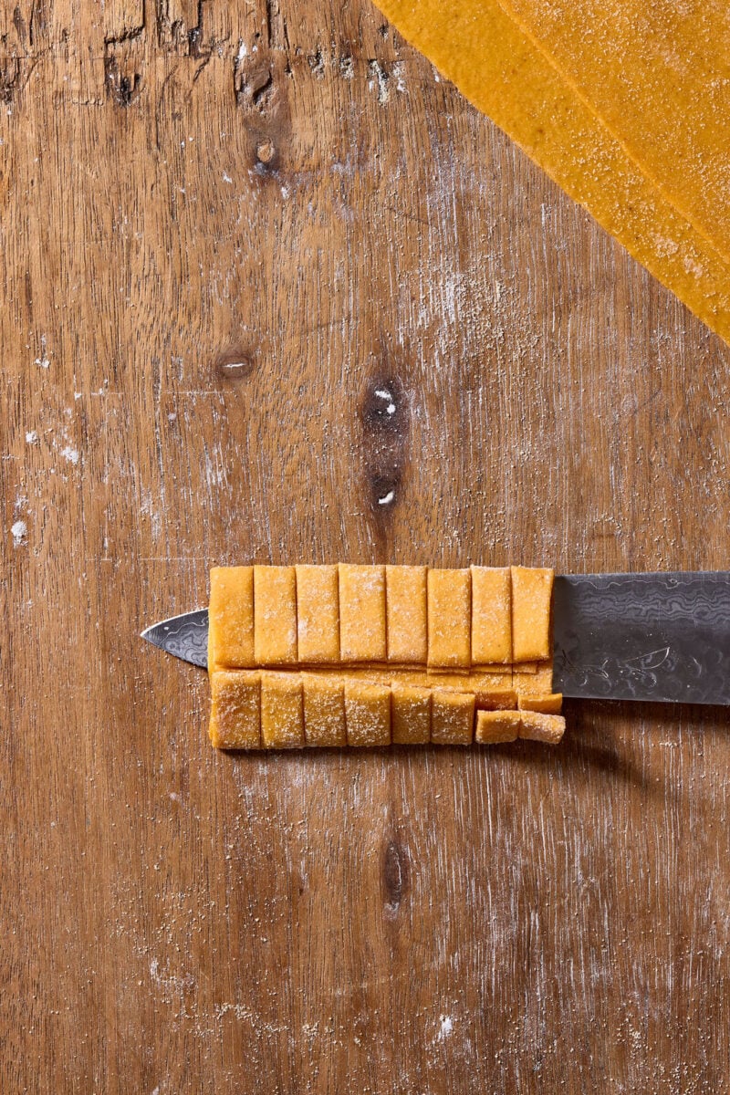 A knife blade holds a stack of fresh, square-cut pasta noodles on a wooden surface dusted with flour.