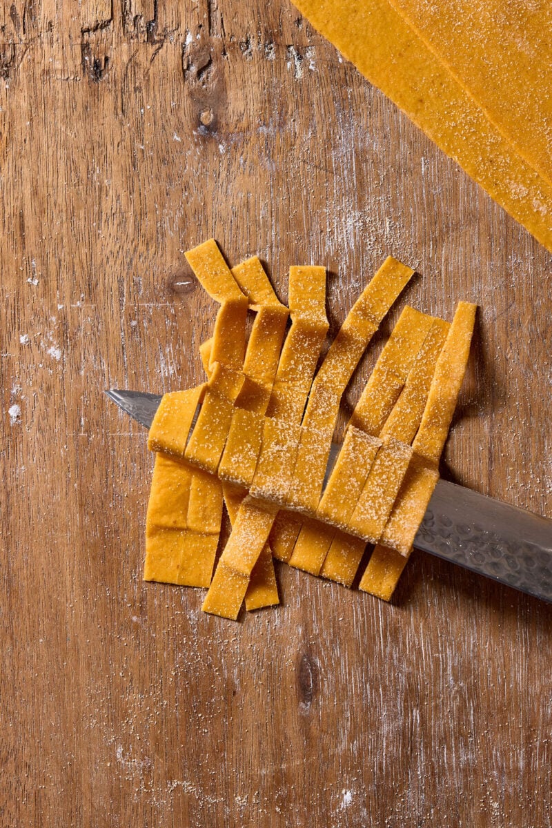 A sharp knife rests horizontally under a pile of freshly cut, orange-hued sourdough pumpkin pasta tagliatelle on a wooden cutting board dusted with flour.