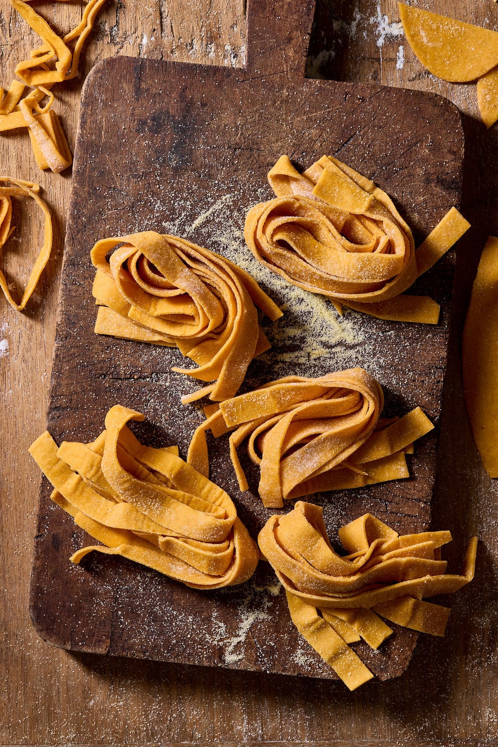Four nests of fresh, golden-hued sourdough pumpkin pasta noodles dusted with flour on a wooden cutting board.