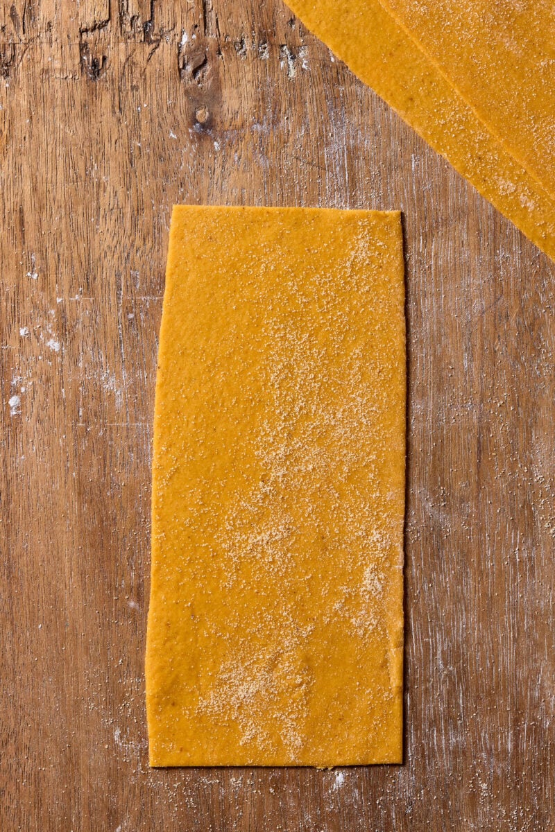 A rectangular sheet of fresh, orange-hued sourdough pumpkin pasta dough dusted with flour on a wooden surface.