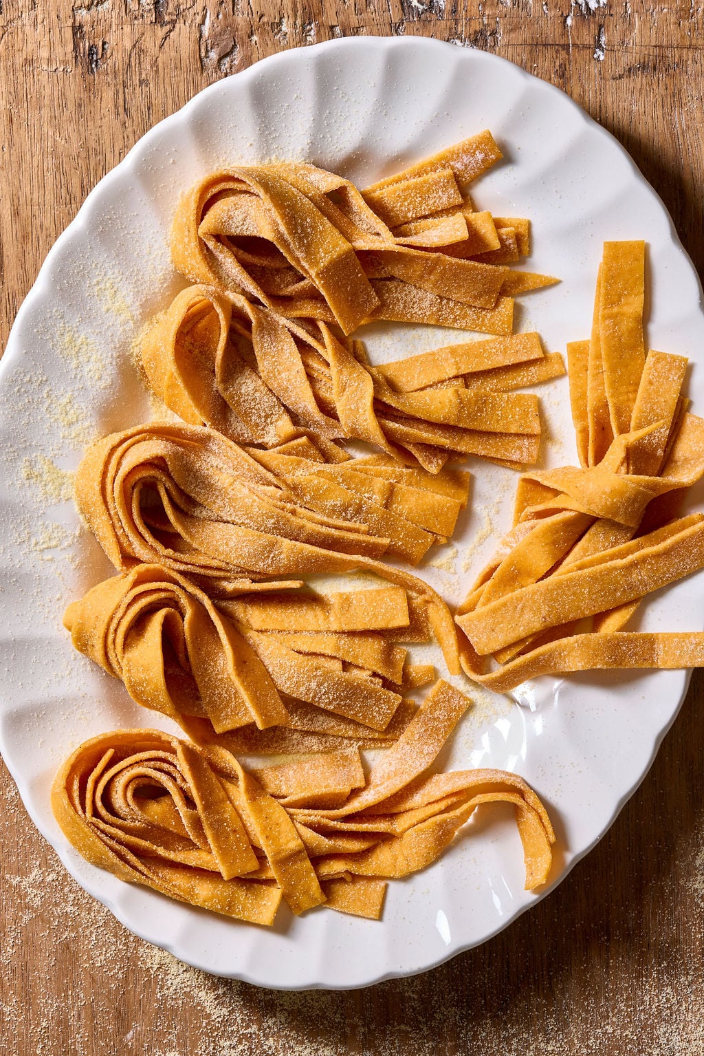Several nests of fresh, flat, sourdough pumpkin pasta noodles dusted with flour on a white scalloped plate.
