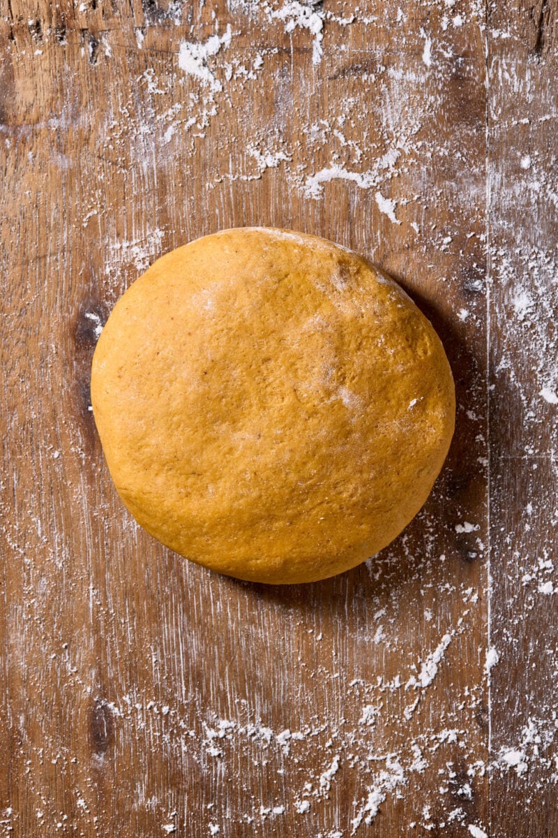 A ball of sourdough pumpkin pasta dough sits on a wooden surface dusted with flour.