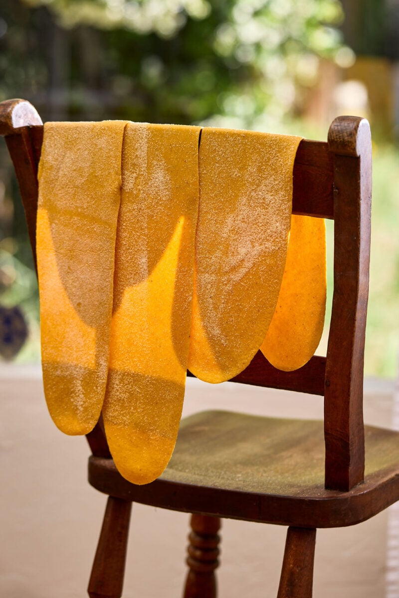 Three sheets of fresh, golden, sourdough pumpkin pasta draped over the back of a wooden chair.