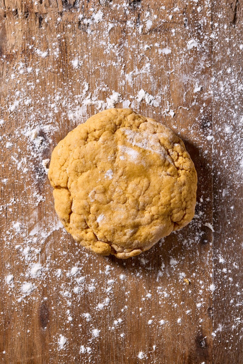 A rough ball of sourdough pumpkin pasta dough on a wooden surface dusted with flour.