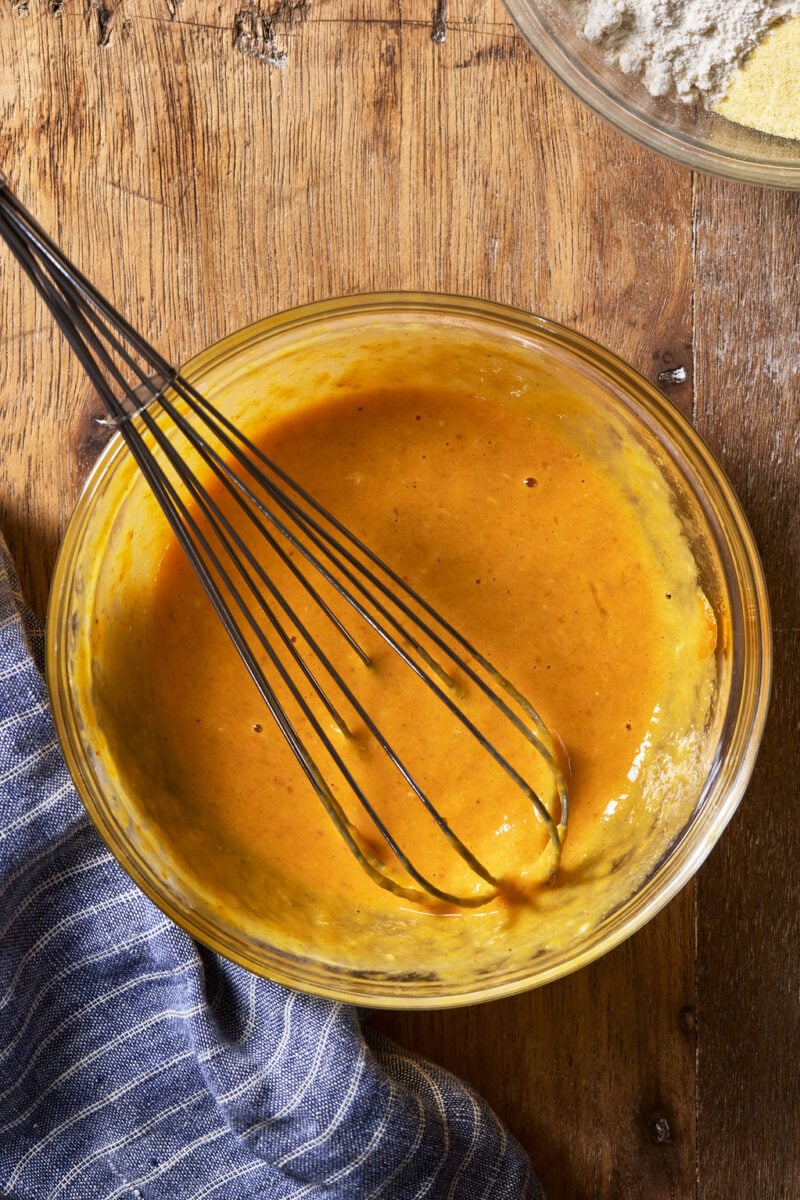 A wire whisk in a glass bowl mixing golden-orange pumpkin mixture on a wooden surface.